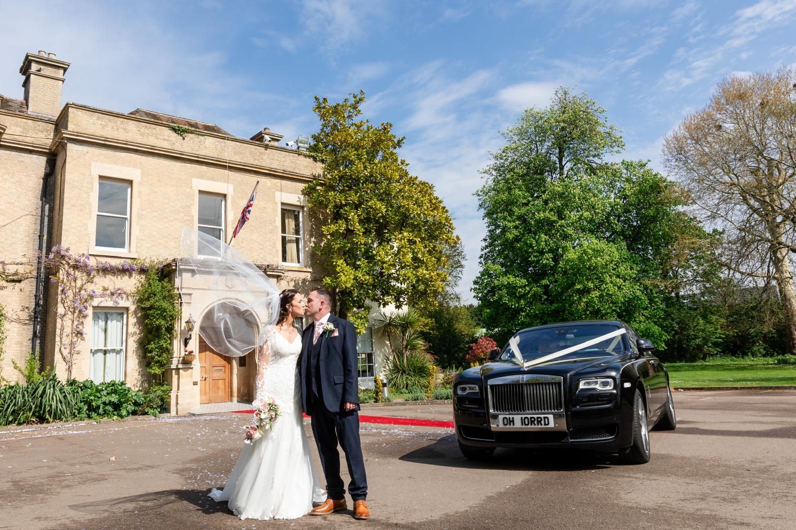 Bride and groom kissing outside country house with Rolls-Royce Ghost